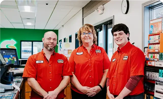 three gas station employees in uniform smiling at the camera from behind the counter
