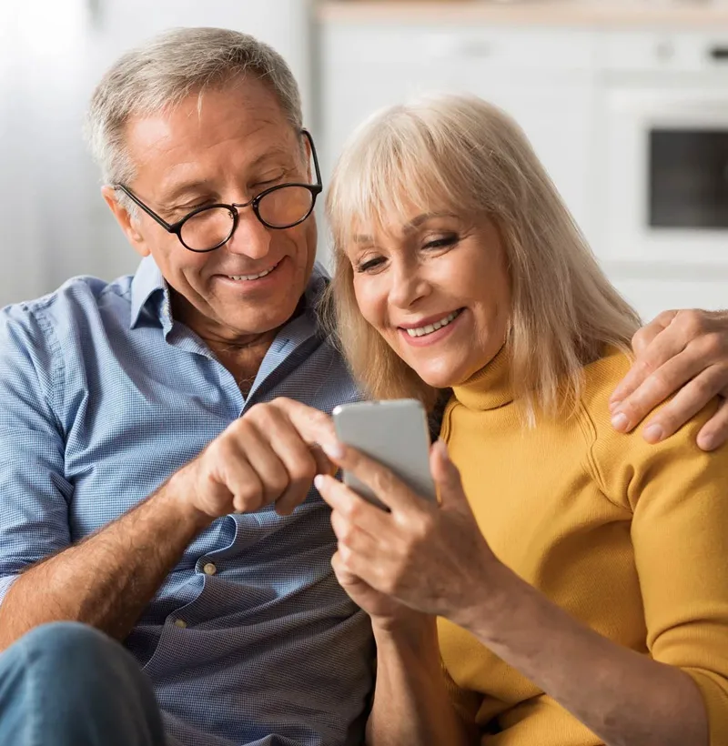 elderly couple look at a phone together while smiling and pointing out details about what they're reading